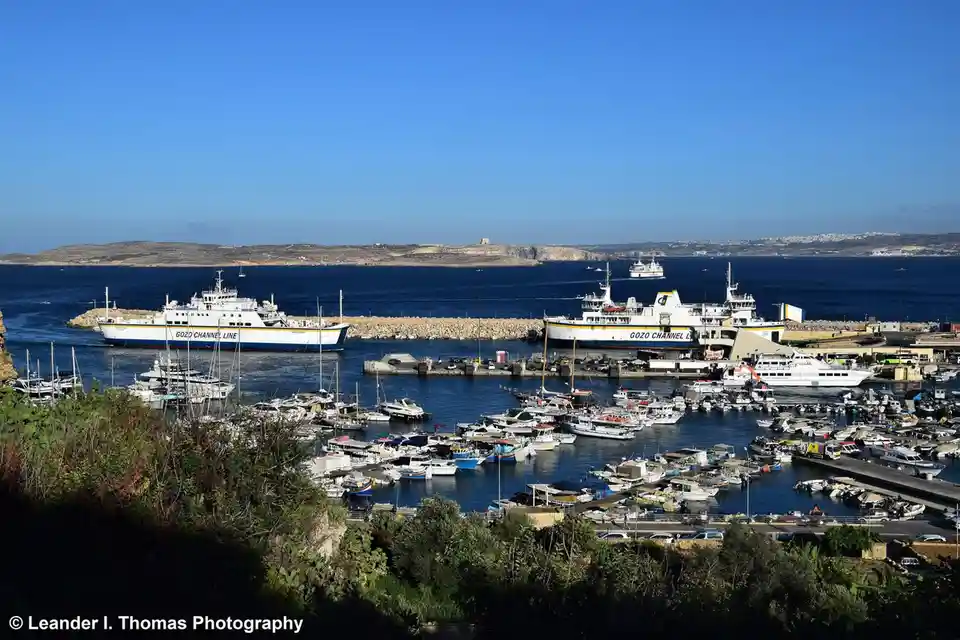 A scenic harbor view with numerous boats and two large ferries labeled 'GOZO CHANNEL LINE' docked at a pier under a clear…