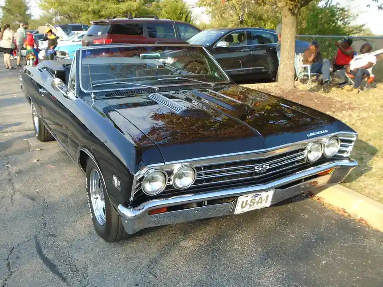 A black 1960s Chevrolet Chevelle SS convertible is parked at an outdoor car show, its shiny hood reflecting trees. People…