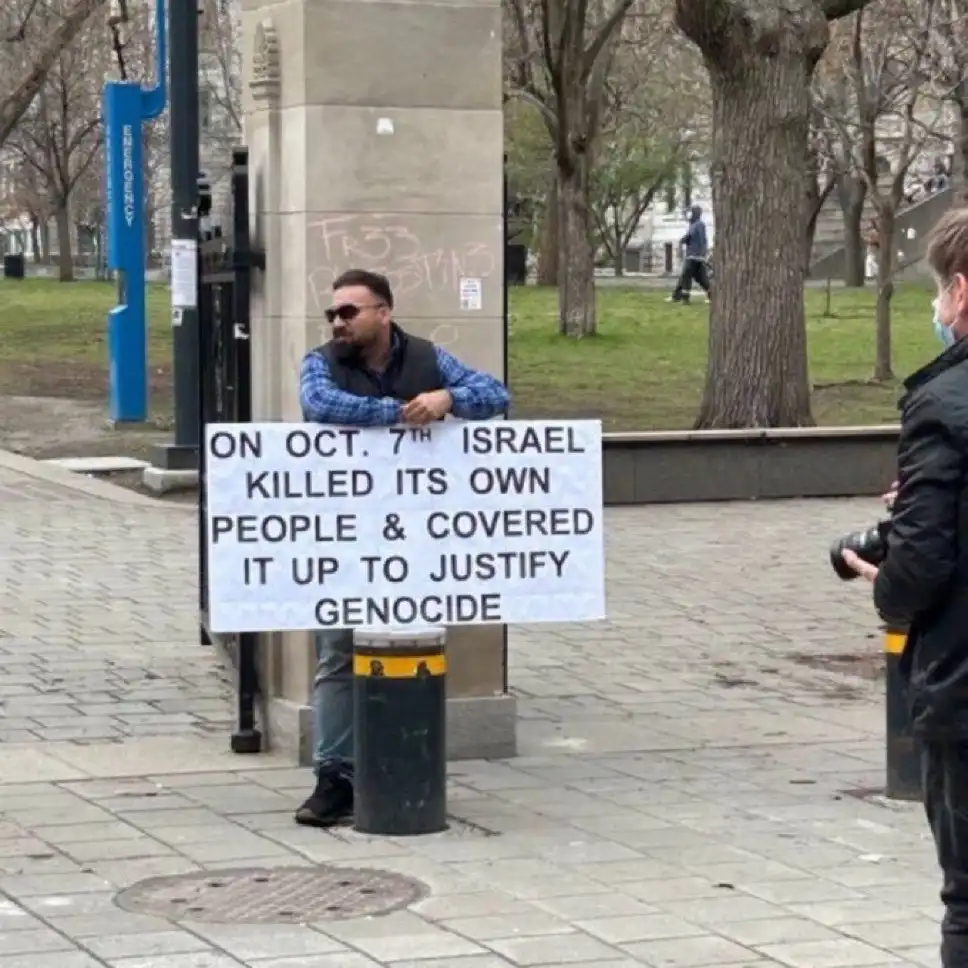 A man wearing sunglasses and a vest stands outdoors holding a white protest sign with black text. The sign reads: 'ON OCT…