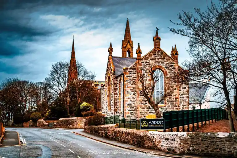 A scenic photo of a stone church with multiple spires under a dramatic cloudy sky, with a sign in the foreground reading…