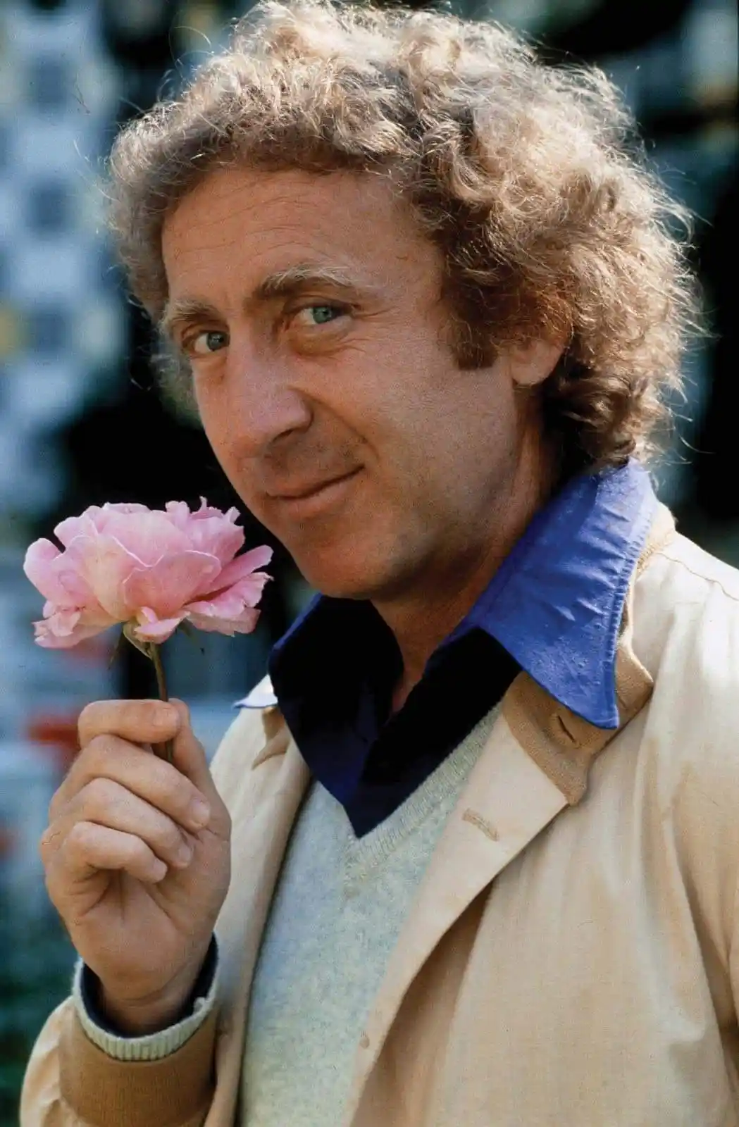 A man with curly brown hair and blue eyes, wearing a beige jacket over a blue collared shirt and light gray sweater, holds a…