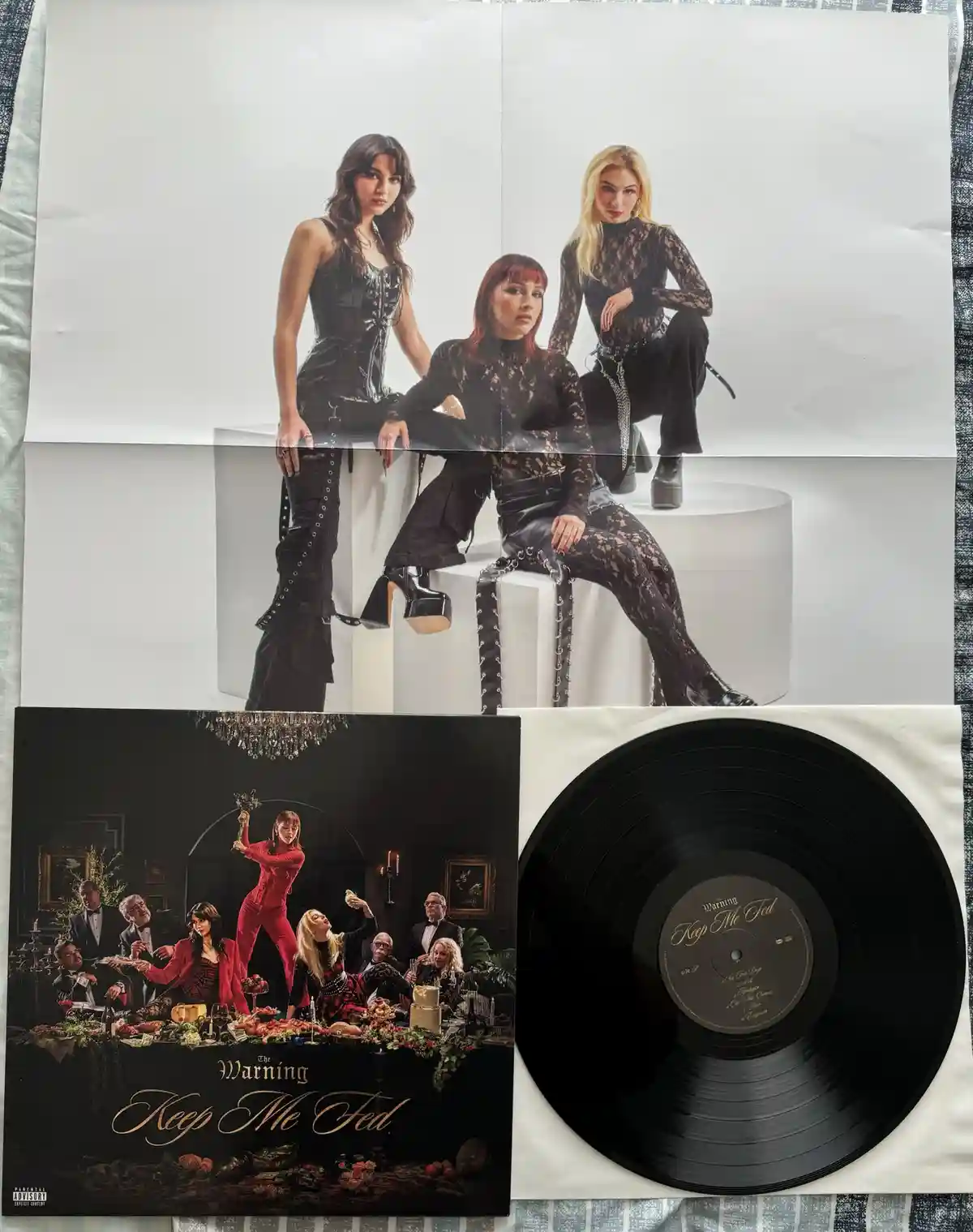 A vinyl record and album cover for 'Keep Me Fed' by The Warning are displayed alongside a large poster featuring the three…