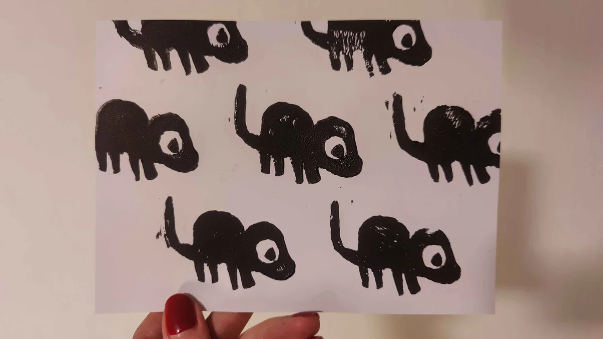 A hand with red nail polish holds up a white paper displaying multiple crude, black-painted elephant silhouettes with large…
