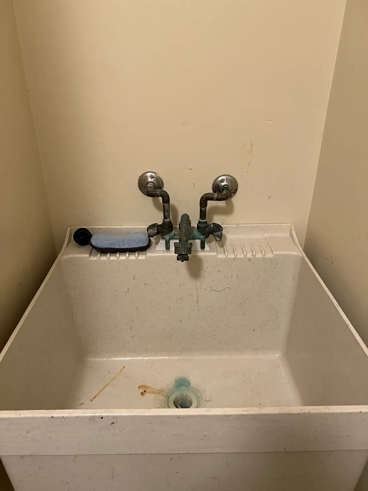 A close-up photo shows a very old, beige, speckled utility sink with a heavily corroded, greenish-bronze faucet and two…