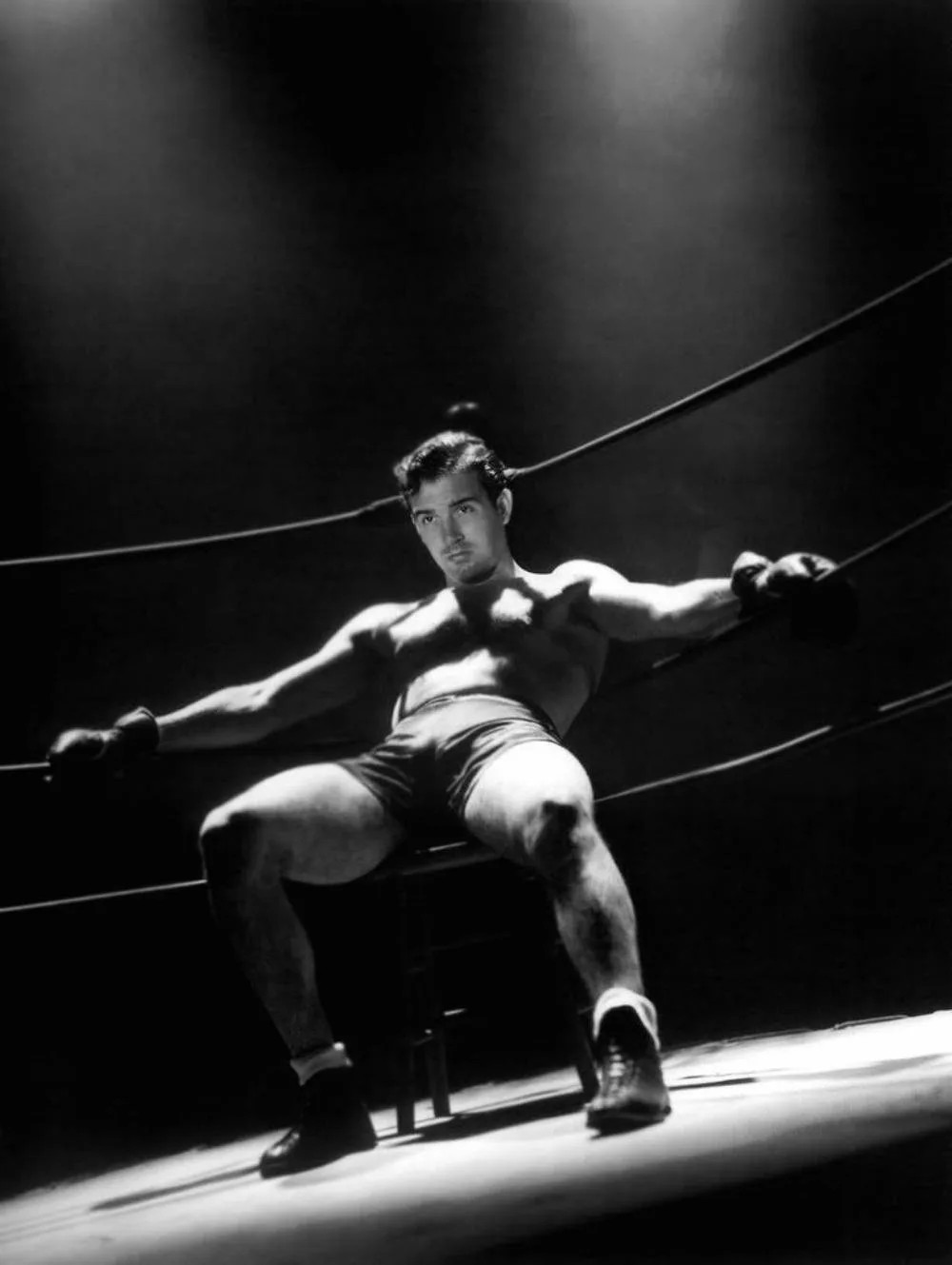 a black and white photo of a muscular man in boxing shorts and gloves sitting on a stool inside a boxing ring, holding the…
