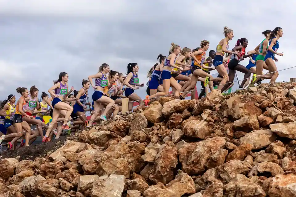 A group of female athletes in running gear are competing in a cross-country race over a rocky terrain under an overcast sky…