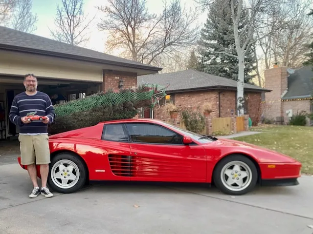 A man stands in a driveway holding a small red toy car, next to a large red Ferrari sports car. The background shows a…