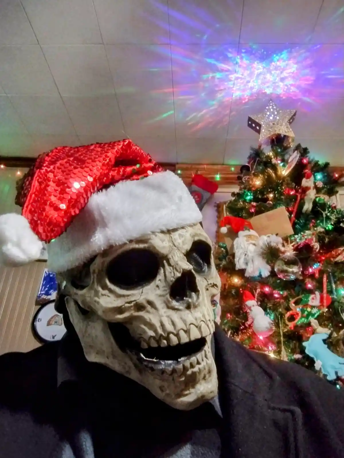 A person wearing a realistic skull mask and a red sequined Santa hat poses in front of a brightly decorated Christmas tree…