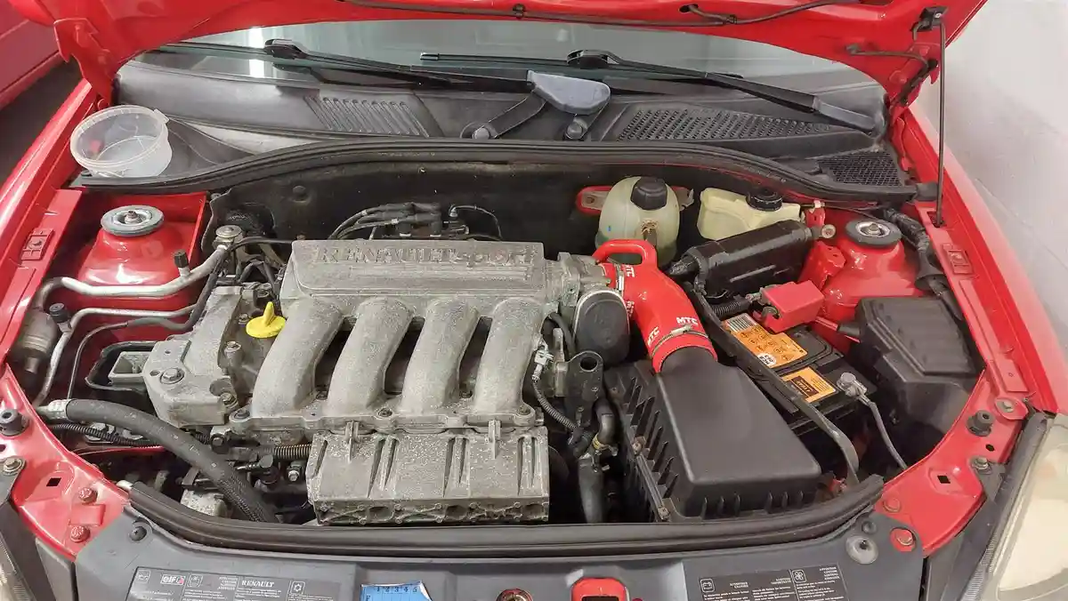 A red car's open hood reveals a dusty engine bay with a prominent 'RENAULT SPORT' engine cover. A red MTS air intake tube is…