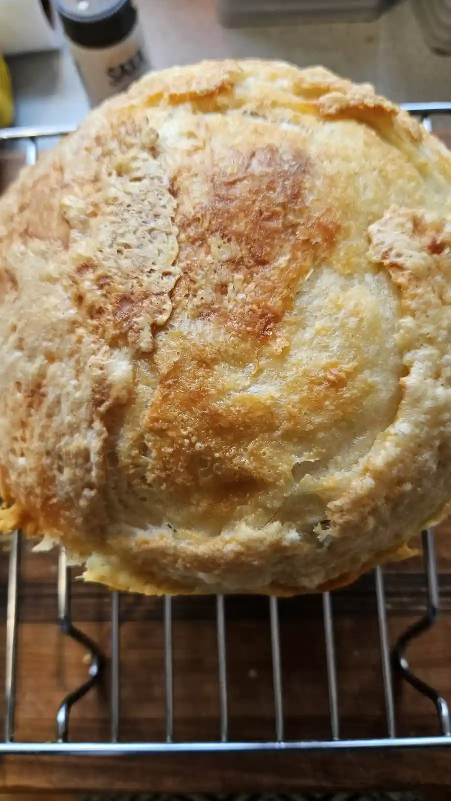 A close-up photo shows a round, golden-brown loaf of bread resting on a metal cooling rack. The bread has a rustic, uneven…
