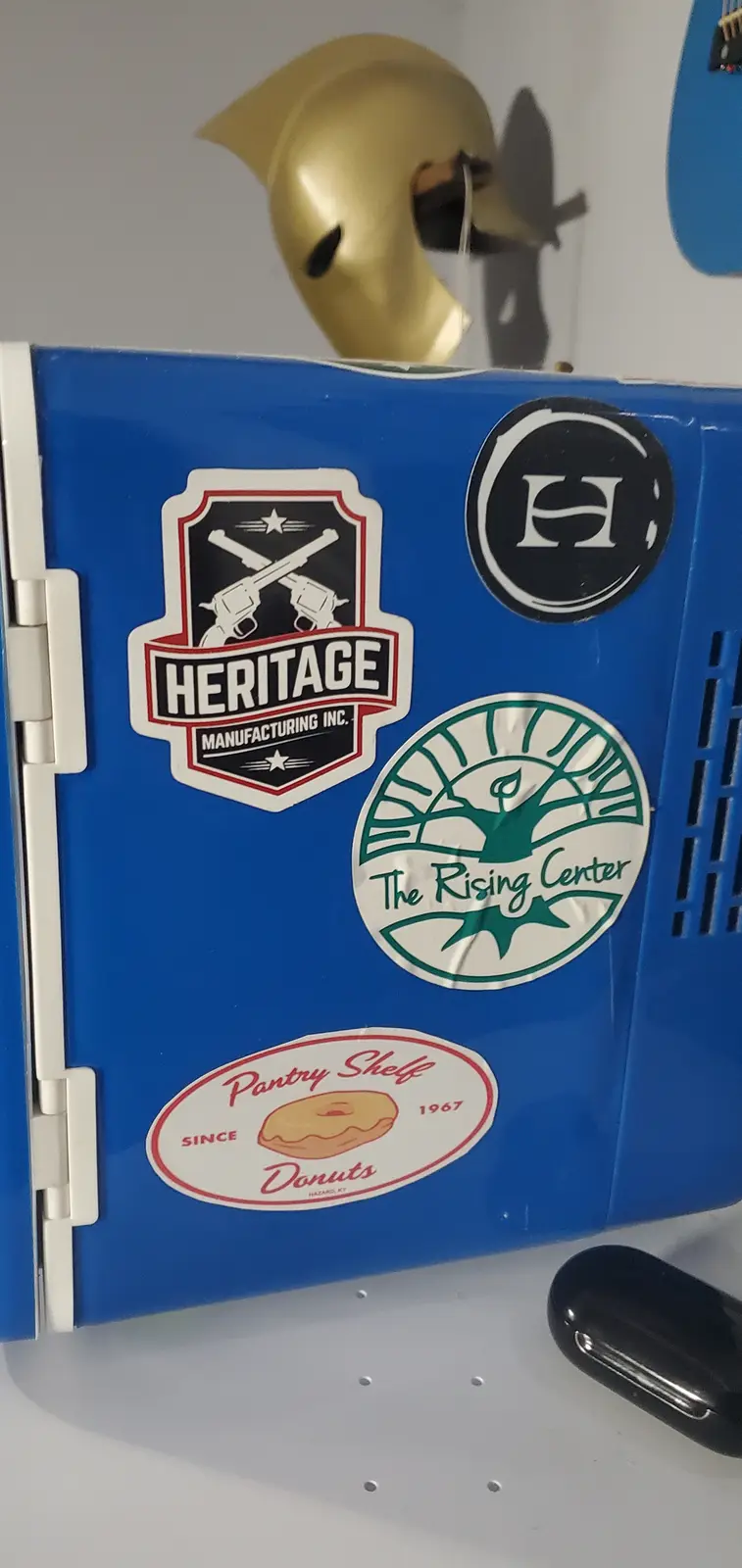 A blue plastic cooler adorned with three stickers: a 'Heritage Manufacturing Inc.' logo with crossed revolvers, 'The Rising…