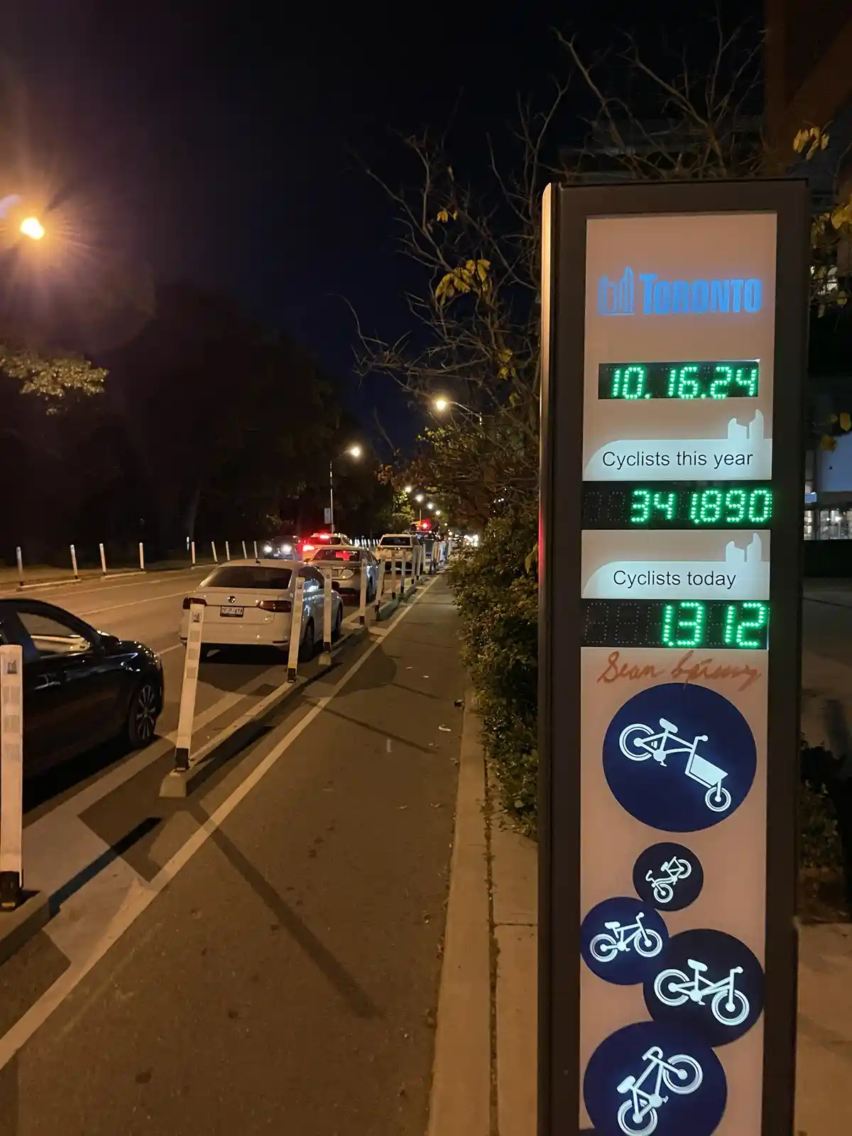 A nighttime photo of a Toronto bike counter displaying 'Cyclists this year: 34 1890' and 'Cyclists today: 13 12', with cars…