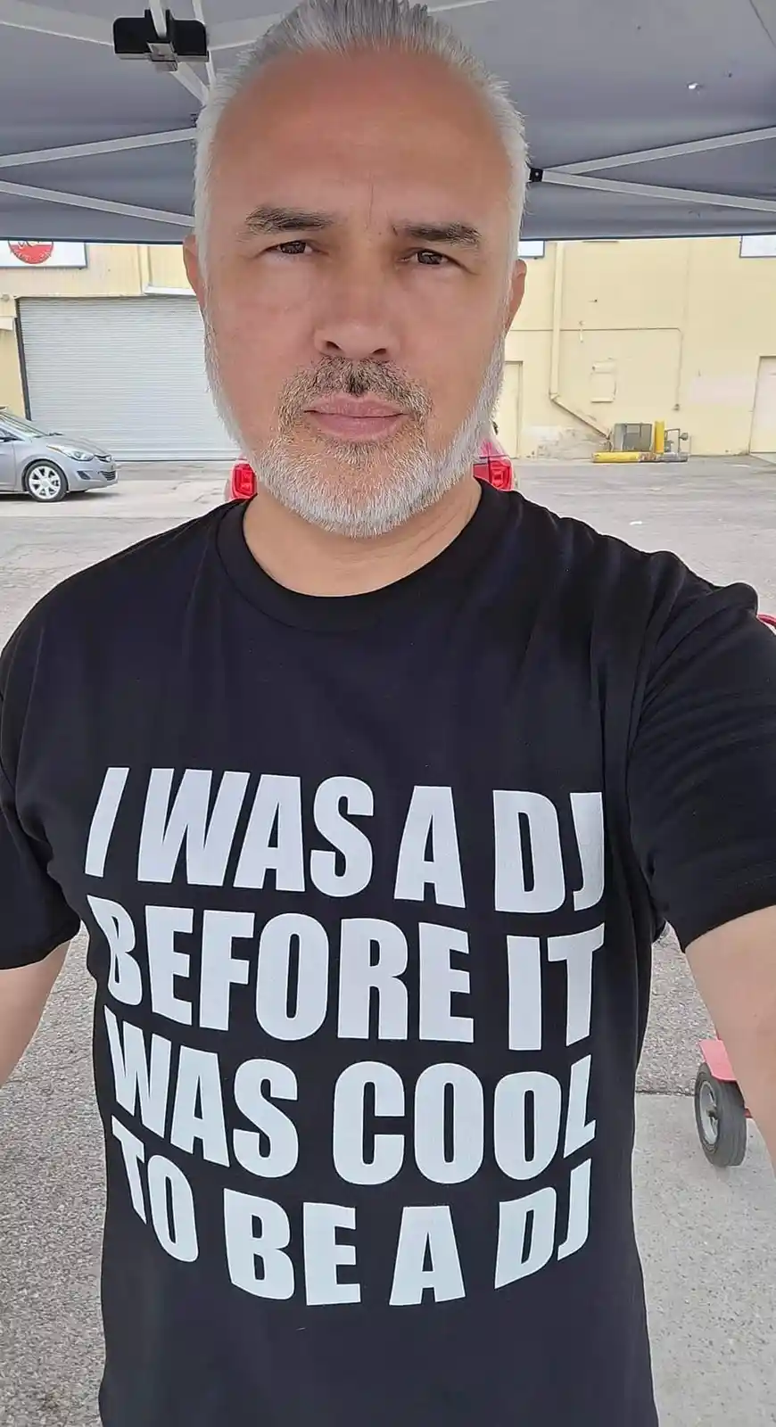 A middle-aged man with gray hair and a beard takes a selfie under a canopy, wearing a black t-shirt with bold white text…