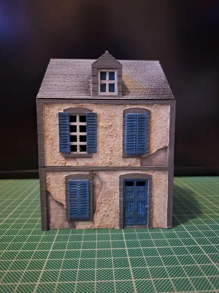 A small, detailed model of a two-story house with blue shutters and a gray roof sits on a green grid mat. The house appears…