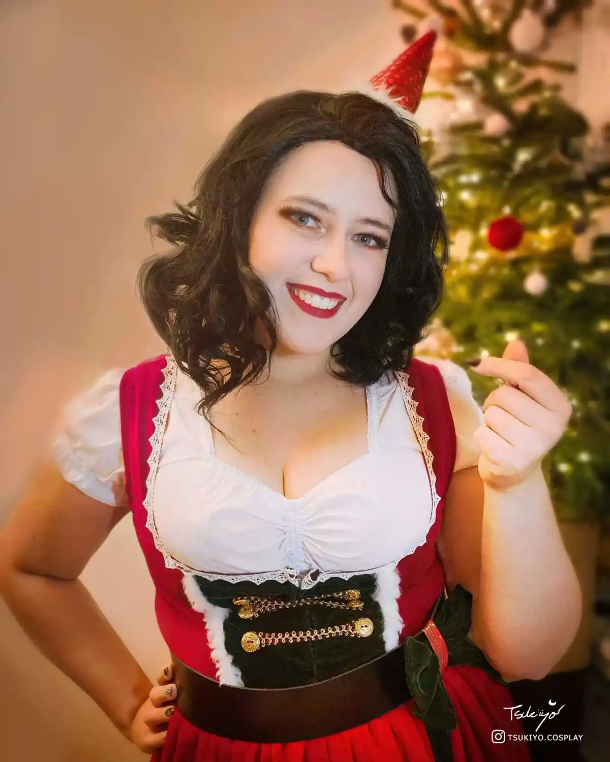 A woman with dark curly hair and red lipstick smiles at the camera while wearing a red and white festive outfit with a black…