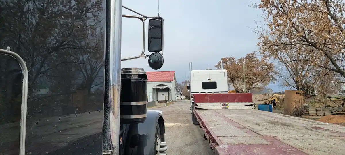 A black semi-truck's side mirror reflects a rural scene with trees and a building, while a white flatbed truck is parked…