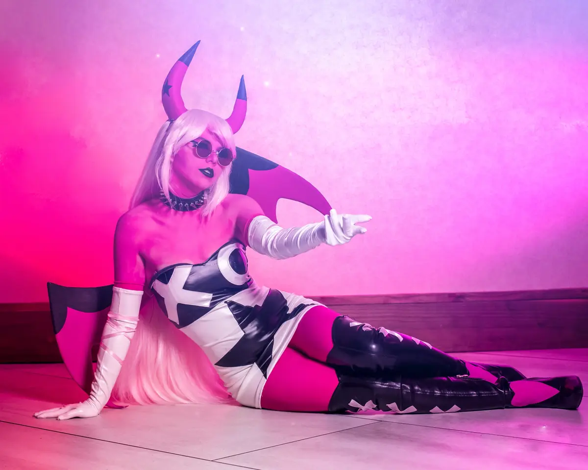 A person in a cosplay of a demonic character with pink horns, white gloves, and a black-and-white outfit is posing on the…