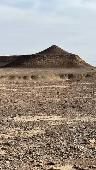 A barren, rocky desert landscape features a large, smooth, cone-shaped mound in the distance under a pale sky. The…