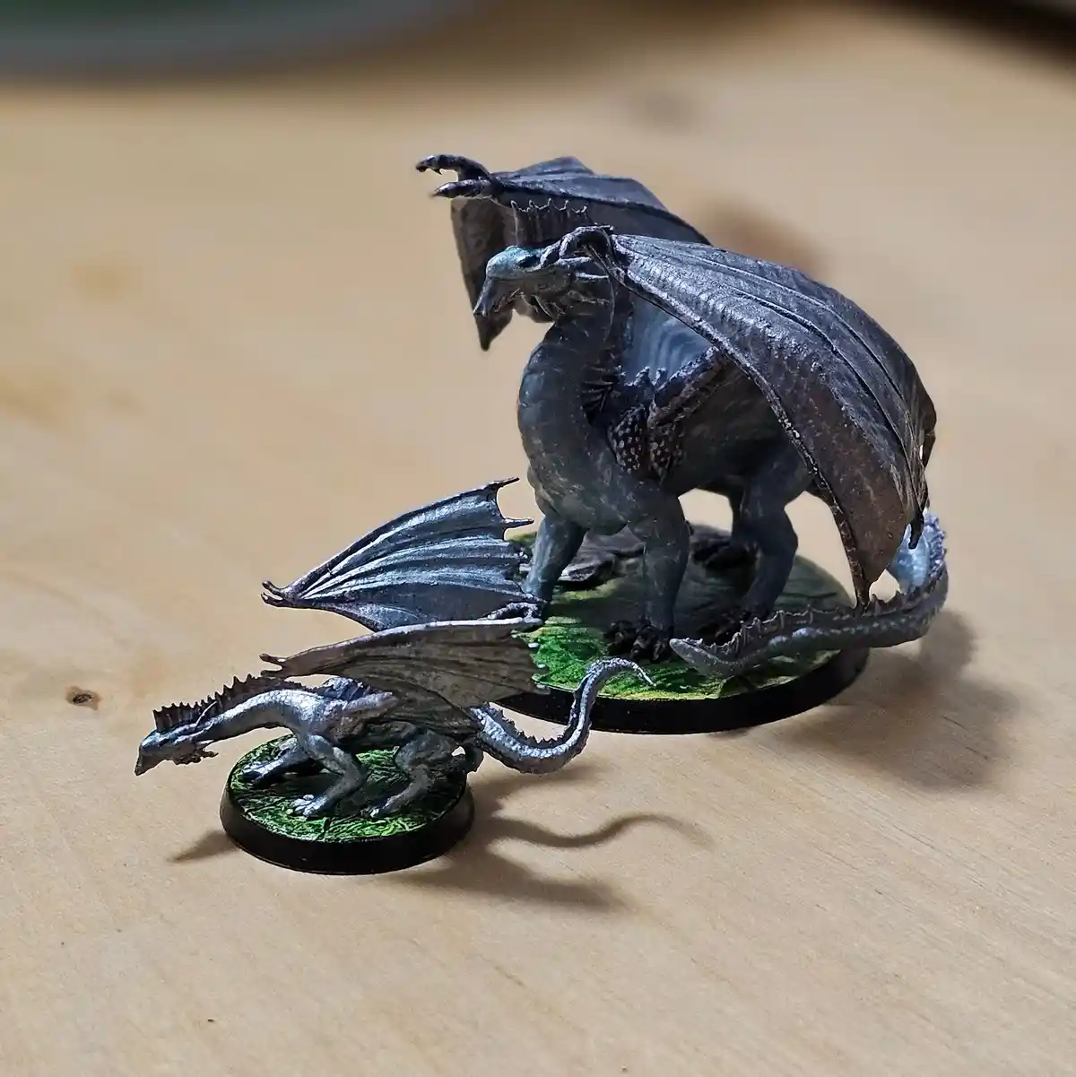 Two painted miniature dragons sit on separate circular bases on a wooden surface. The larger, darker dragon stands upright…