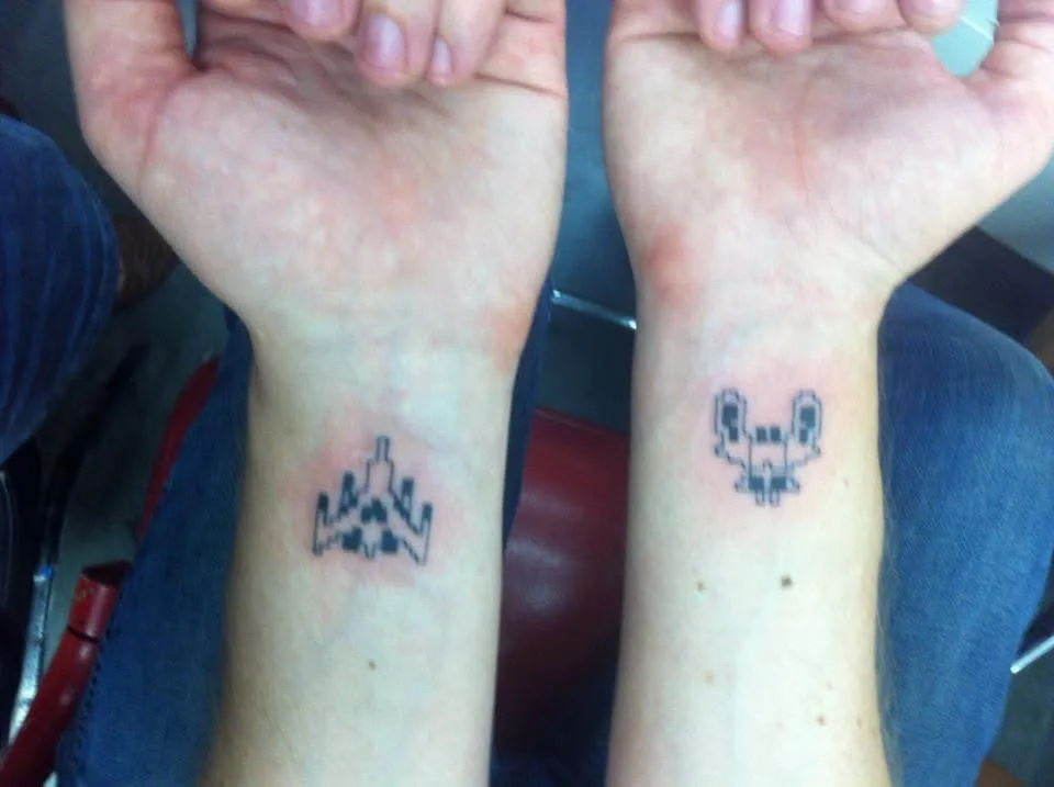 Two wrists are shown side-by-side, each with a small black ink tattoo. The left wrist has a pixelated spaceship design, and…