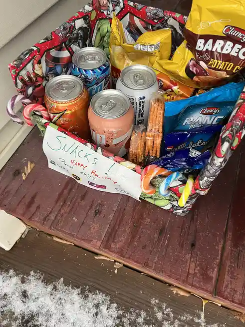 A basket filled with assorted snacks including potato chips, pretzels, candy canes, and several soda cans sits on a wooden…