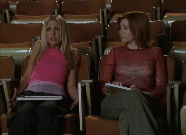 Two women sit in an auditorium with brown seats; the blonde woman on the left looks shocked and dismayed, while the redhead…