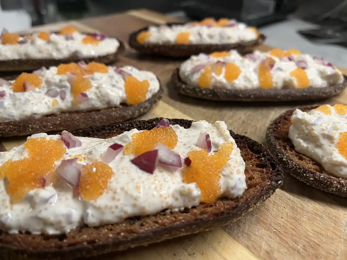 Several dark rye bread slices are arranged on a wooden cutting board, each topped with a creamy white spread, bright orange…
