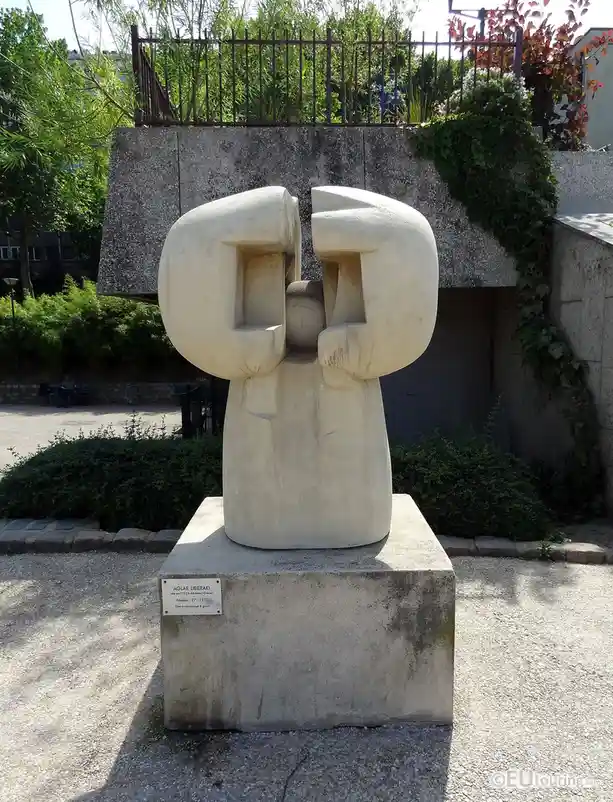 A photograph of a large, abstract white stone sculpture of two figures embracing, standing on a concrete pedestal in a park…