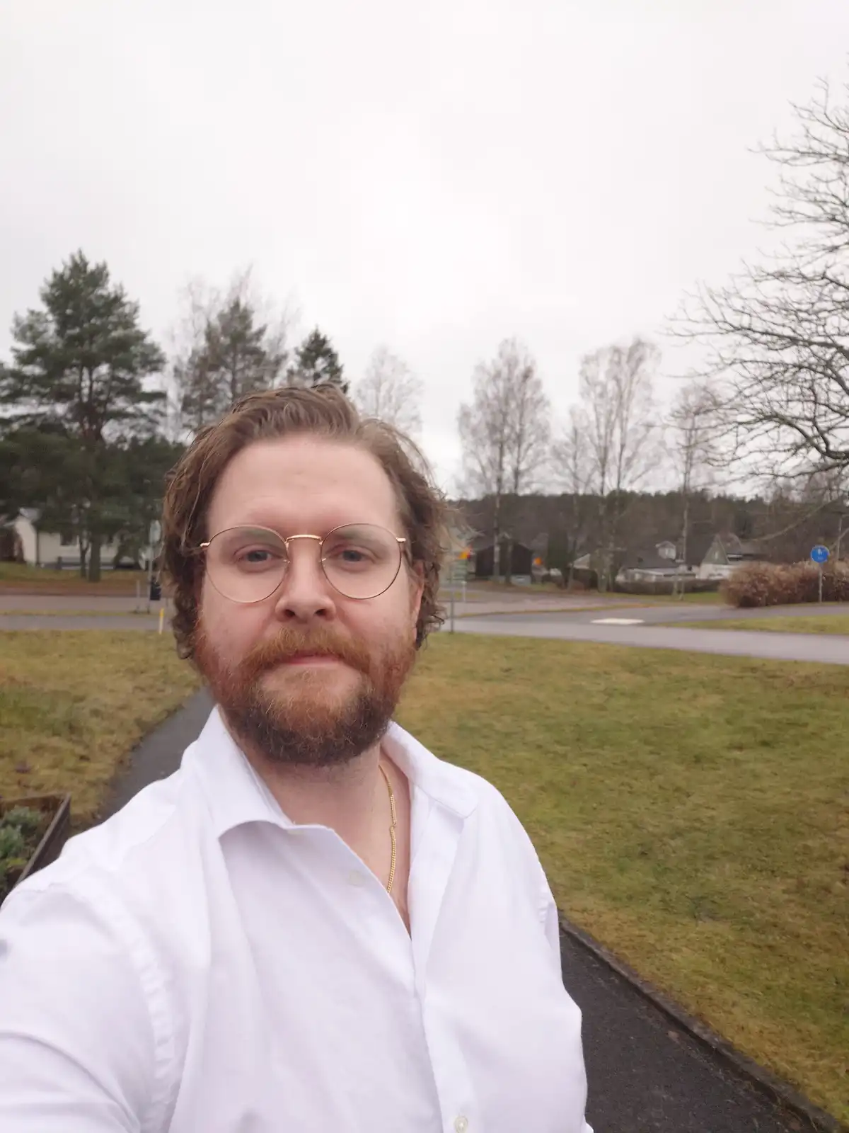 A man with a beard and glasses takes a selfie outdoors on an overcast day, wearing a white shirt and a gold chain. The…