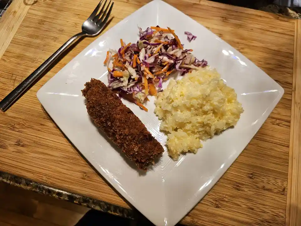 A white square plate holds a breaded sausage, a scoop of yellow rice, and a side of coleslaw with purple cabbage and…