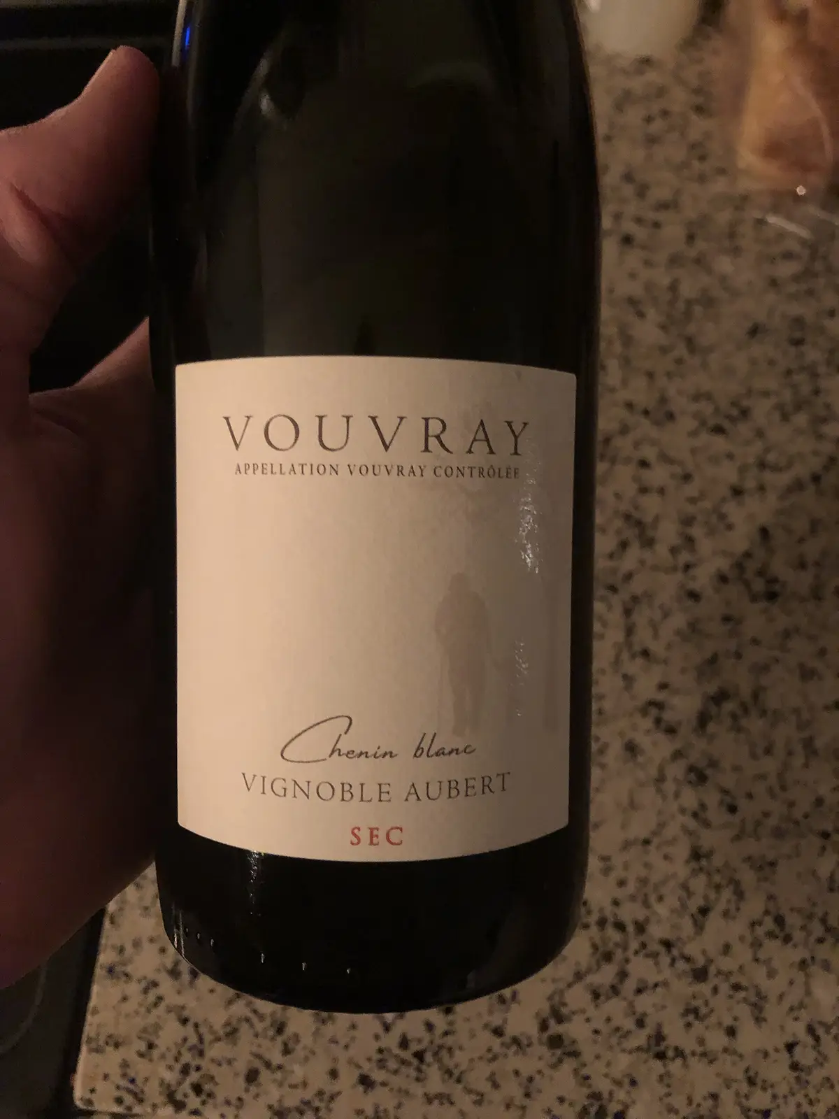 A hand holds a dark wine bottle with a white label reading 'VOUVRAY APPELLATION VOUVRAY CONTRÔLÉE', 'Chenin blanc'…