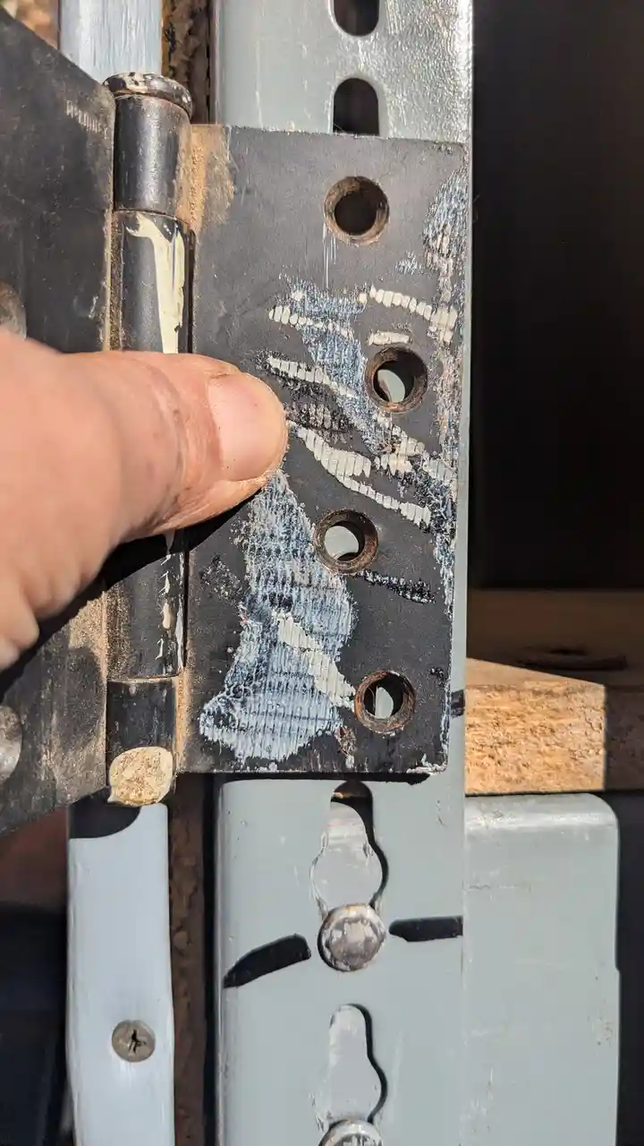 A close-up photo shows a person's thumb pointing at a weathered metal hinge with chipped white paint revealing the dark…