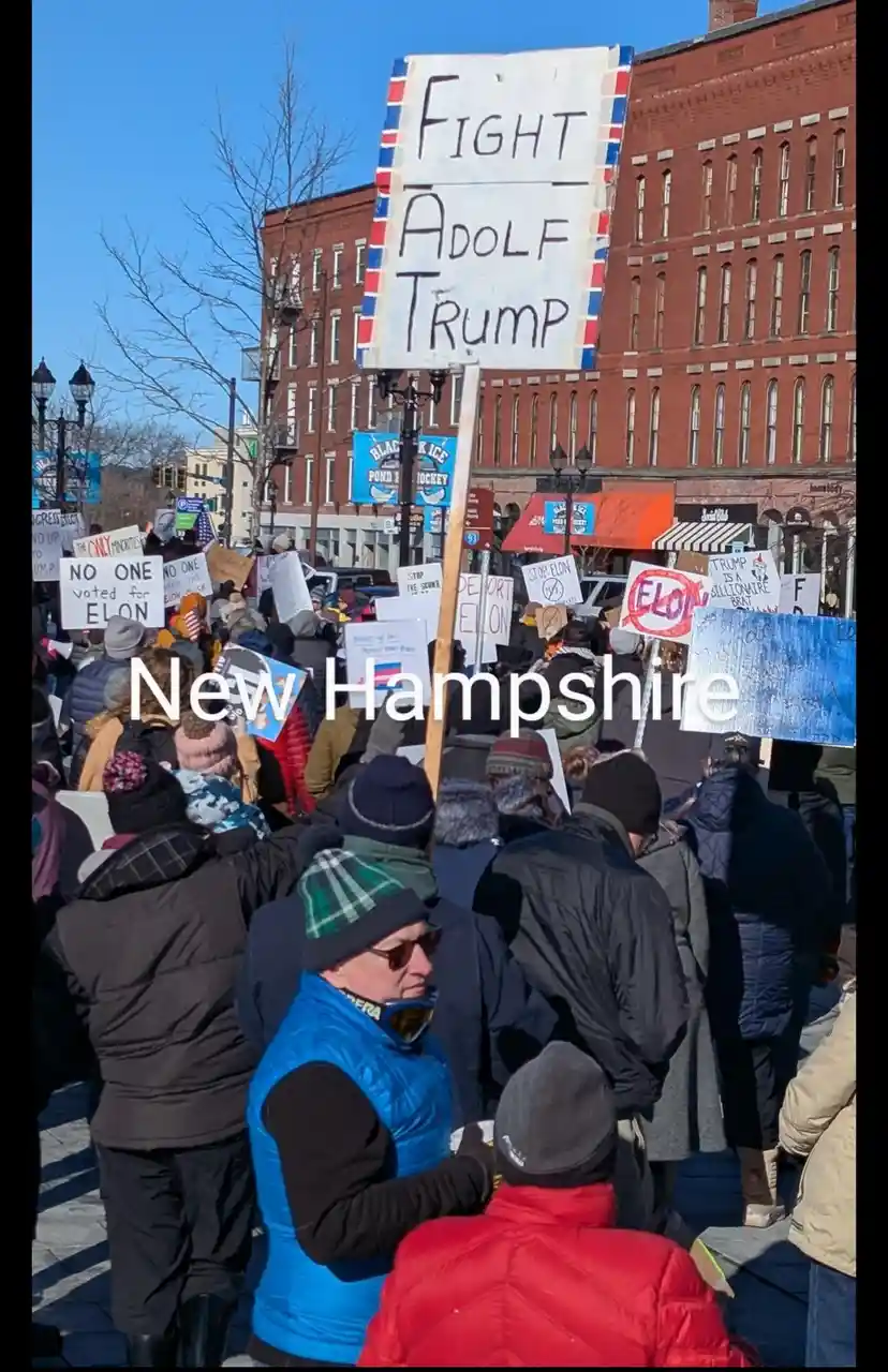 A crowd of protesters in winter coats and hats holds signs at a rally in New Hampshire. A large handmade sign reads 'Fight…