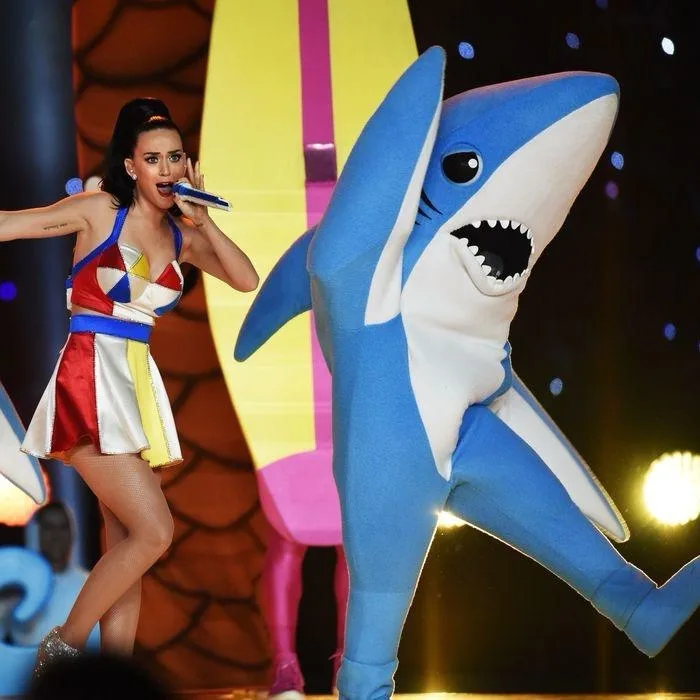 A woman in a colorful geometric outfit sings into a microphone while dancing next to a person in a large blue and white…