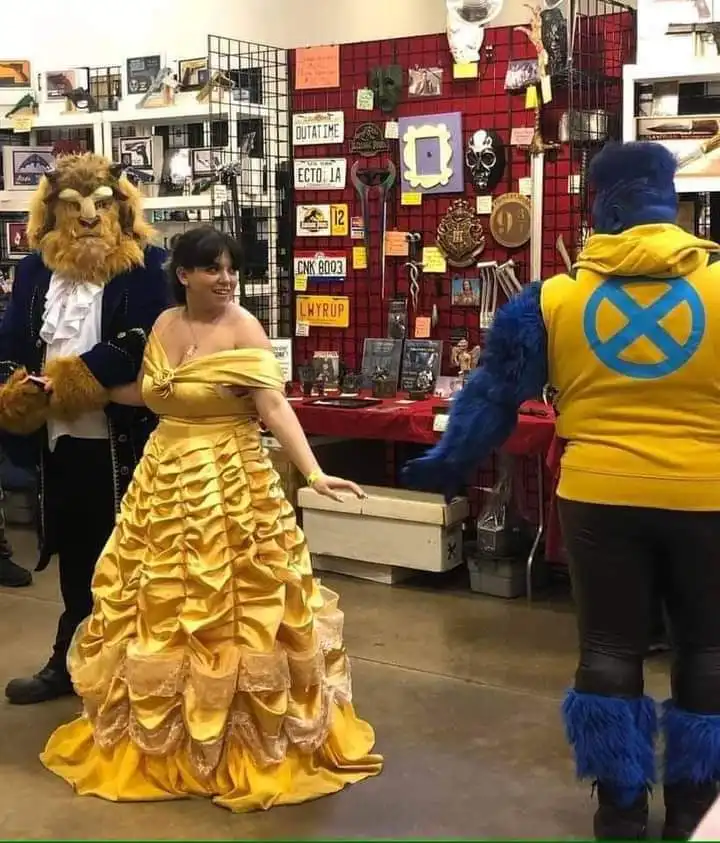 A woman in a yellow Belle costume smiles while standing between a Beast cosplayer in a furry costume and a blue-furred Beast…