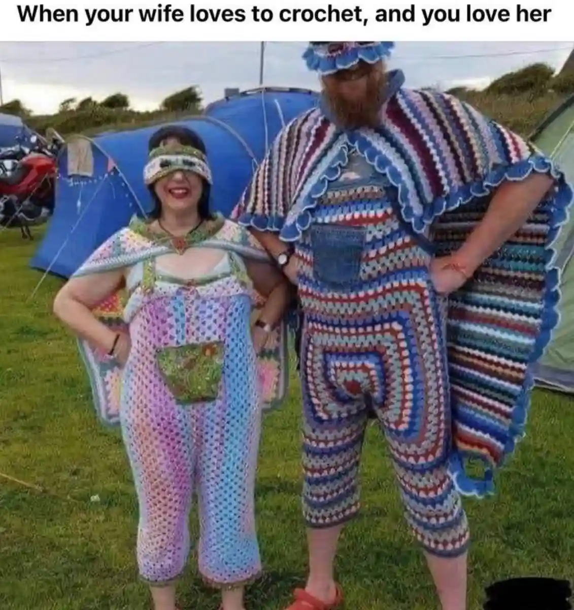 A man and woman stand outdoors on grass near tents, both wearing full-body, colorful, crocheted outfits. The woman wears a…