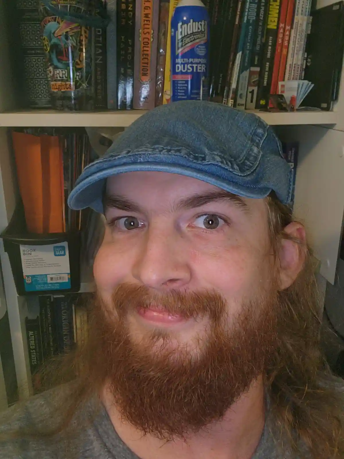 A man with a red beard and a denim cap stares directly at the camera with wide eyes and a slight smirk, standing in front of…