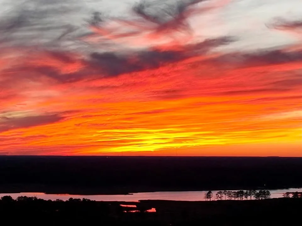 A vibrant sunset with fiery orange and red clouds over a dark landscape featuring a body of water and silhouetted trees. The…