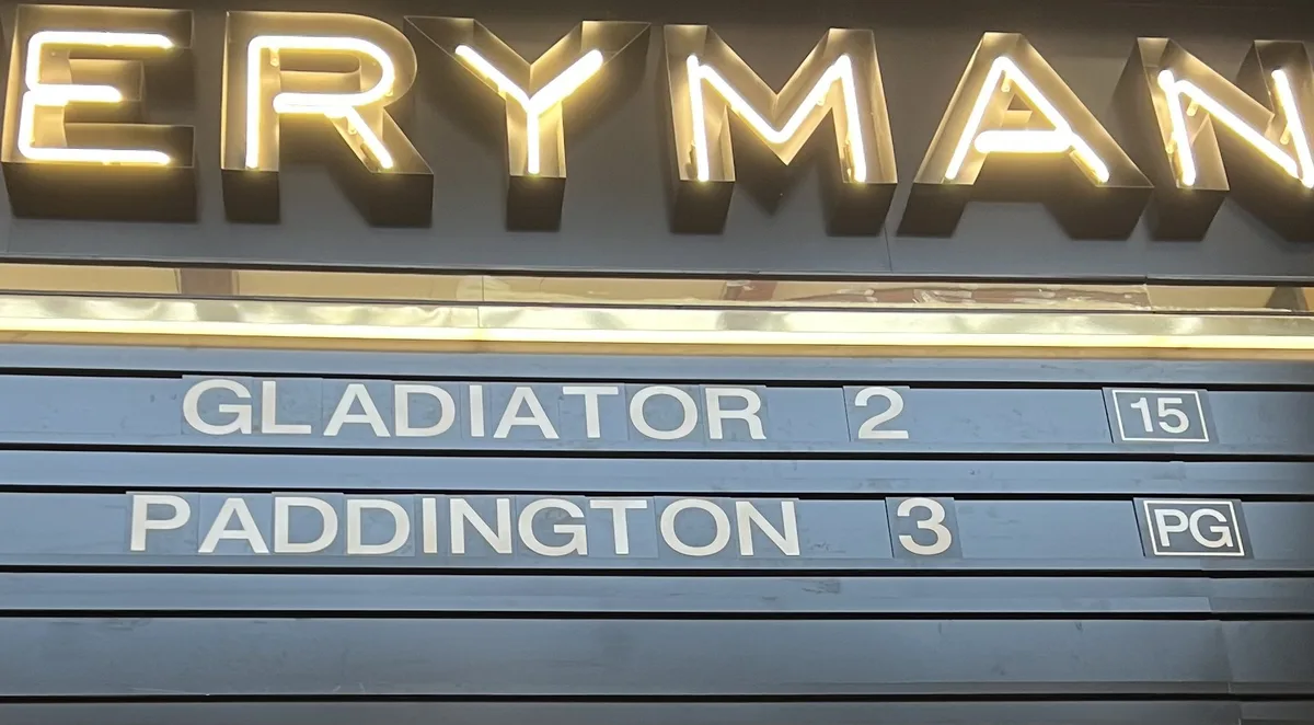 A close-up of a cinema marquee for 'ERYMAN' showing 'GLADIATOR 2' rated 15 and 'PADDINGTON 3' rated PG, creating a humorous…