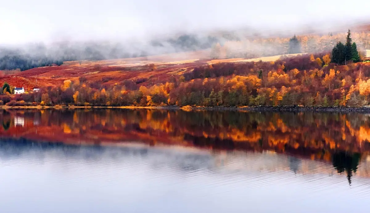 A serene autumn landscape with a calm lake reflecting vibrant orange and red foliage on rolling hills partially obscured by…