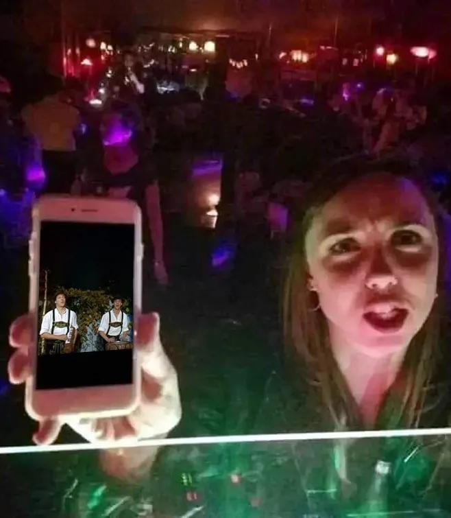 A woman in a dimly lit, crowded club with colorful lights holds up a smartphone displaying a photo of two men in traditional…