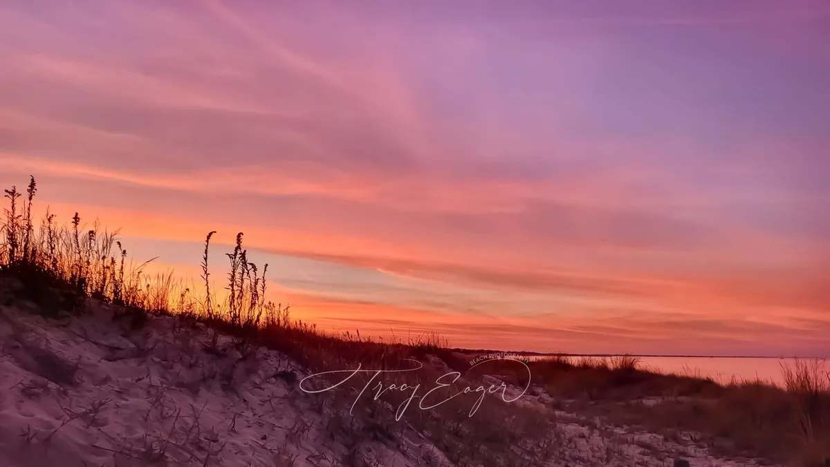 A serene beach sunset photo with vibrant orange and purple skies over sand dunes and grasses, featuring a watermark that…