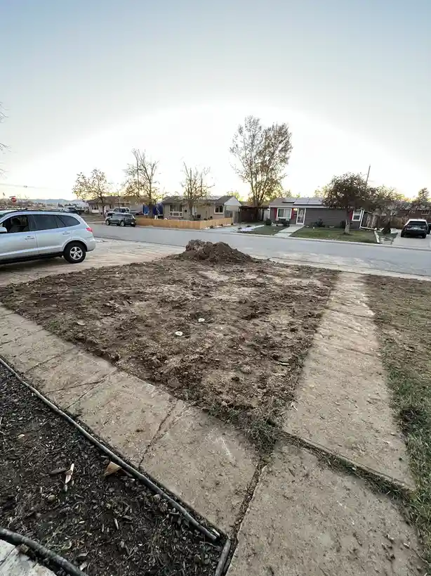 A residential front yard with a large, freshly dug dirt patch where grass used to be, bordered by concrete walkways and a…
