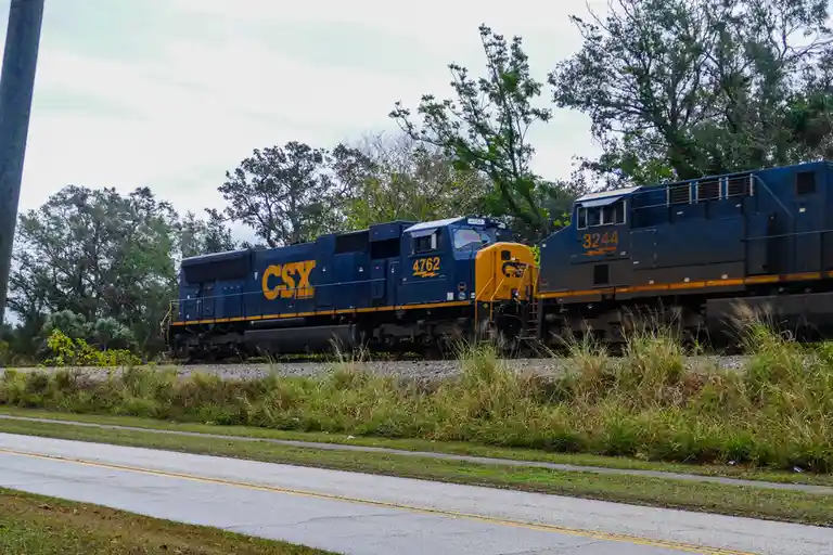 Two blue CSX freight locomotives, numbered 4762 and 3244, are shown side-by-side on parallel tracks near a road with grass…