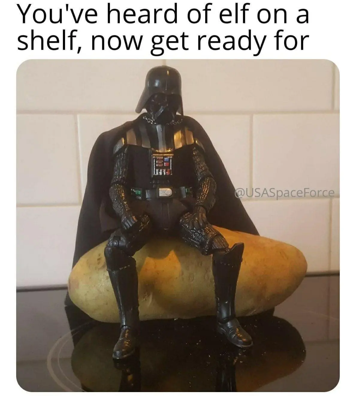 A Darth Vader action figure is seated on a large potato, which rests on a black glass stovetop. The background shows white…