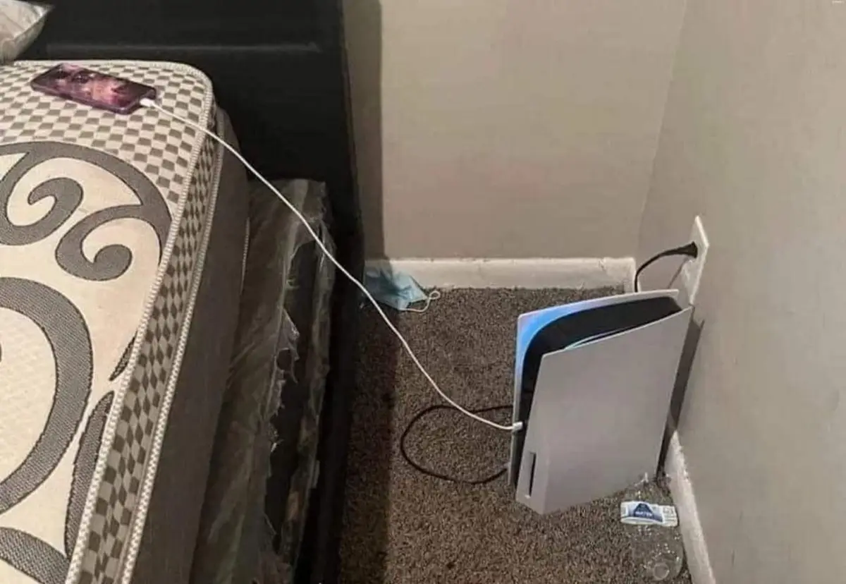 A silver PlayStation 5 console sits upright on a carpeted floor next to a wall outlet, with a white charging cable plugged…