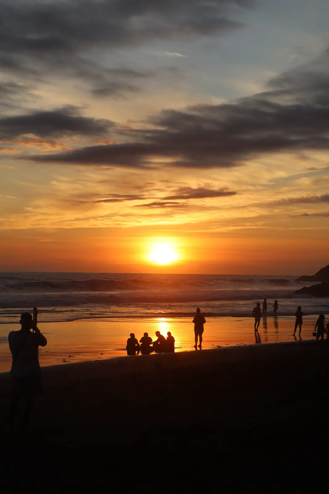 A silhouette of a person holding a phone to take a photo of a sunset on a beach, with other people enjoying the view. The…