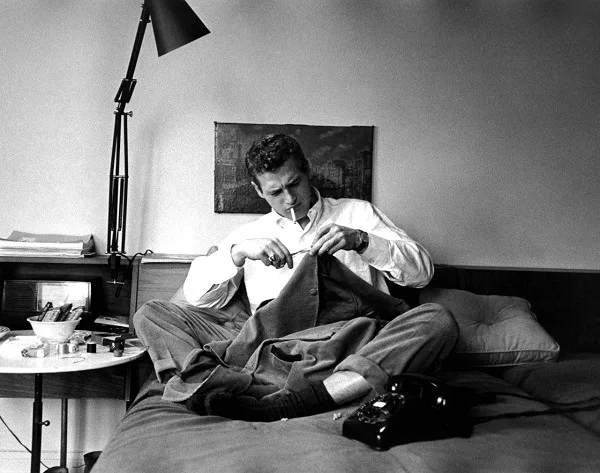 a black and white photo of a man sitting on a bed, smoking a cigarette while holding a jacket. He is surrounded by a bedside…