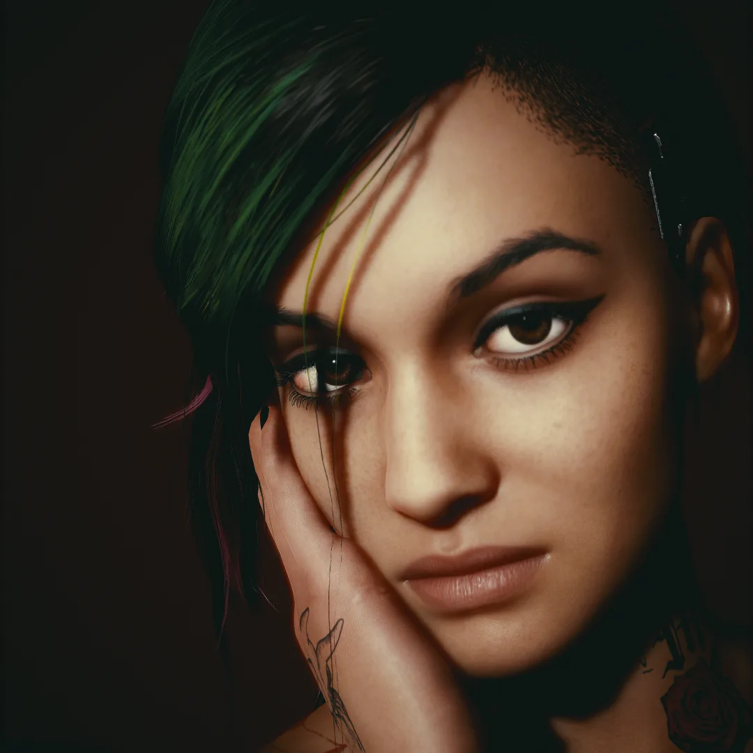 A close-up of a woman with dark hair streaked with green and pink, her hand resting on her cheek. She has tattoos on her…