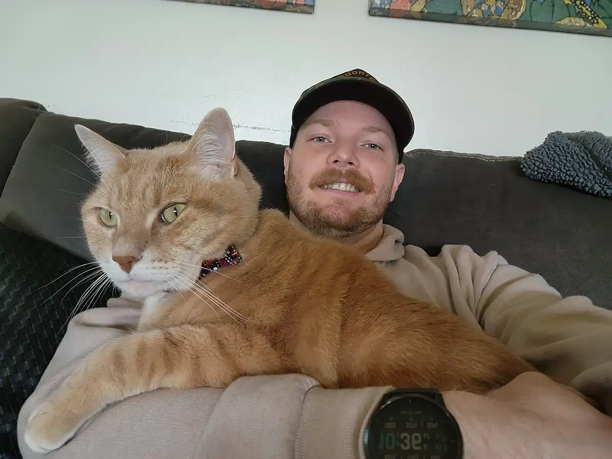 A man with a mustache and cap smiles at the camera while lying on a couch, with a large orange tabby cat resting on his…
